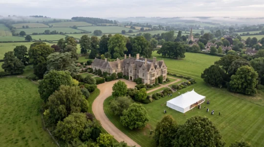 Wide aerial-style editorial photograph of a grand British country estate at golden hour with a wedding marquee visible in the grounds below