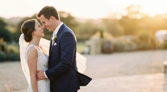 A couple in elegant wedding attire sharing an intimate, laughing moment lit by warm golden-hour sunlight, styled like a high-fashion magazine editorial